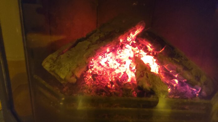 En brinnande vedeld i en öppen spis, glödande eld, varmvit belysning, bakom glas.