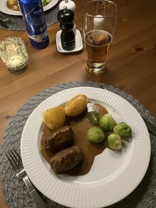 Middagsmåltid med köttbullar, potatis, brysselkål, sås, öl, salt- och pepparkvarnar på träbord.