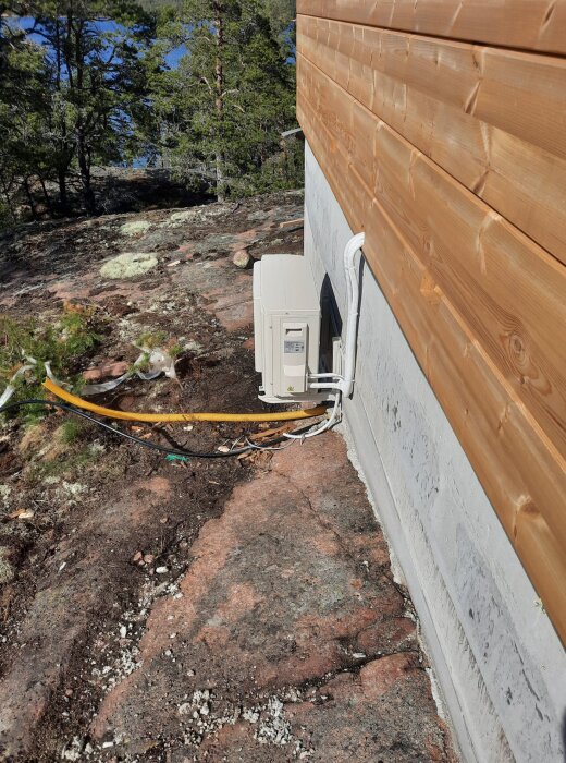 Luftvärmepump monterad på husvägg med synliga kablar, i skogsmiljö med tallar och klippgrund.