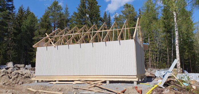 Hus under konstruktion med trästomme, takstolar synliga, i skogsmiljö på en solig dag.