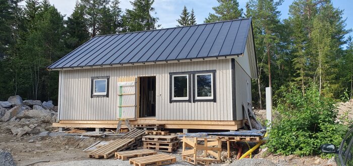 Litet hus under konstruktion, stående på pallar, bland byggmaterial, skog runtomkring.