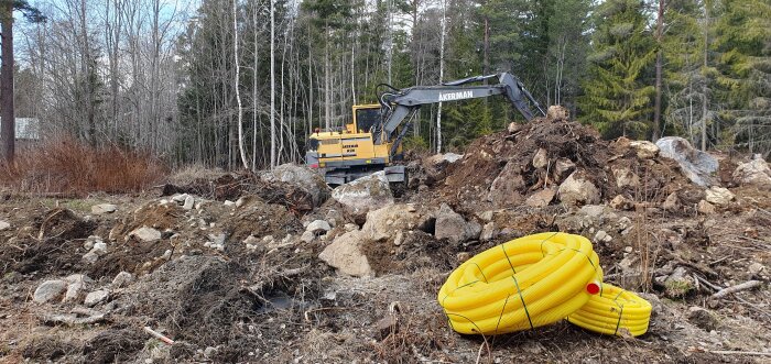 Gul grävmaskin i skogsröjning, stenar, jord, gul rörspole i förgrunden, barr- och lövträd.