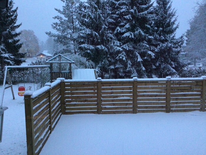 Snötäckt trädgård med staket, lekstuga och gungställning i skymningen. Träd och hus i bakgrunden.