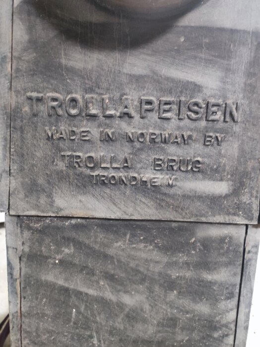 Text på en skulptur: "TROLLAPEISEN MADE IN NORWAY BY TROLLA BRUG TRONDHEIM". Sliten, nött detalj.