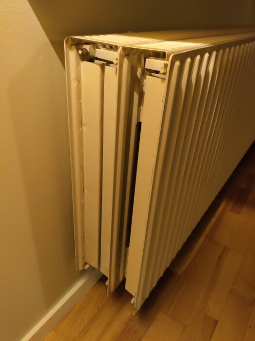 En vit värmeelement/radiator i ett hörn, inomhus, på ett trägolv och intill en vägg.