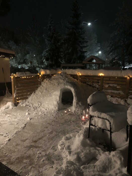 Kväll, nybyggd snöiglo, ljusslingor, snötäckt stol, fotspår i snön, träd, mysig vinterstämning.