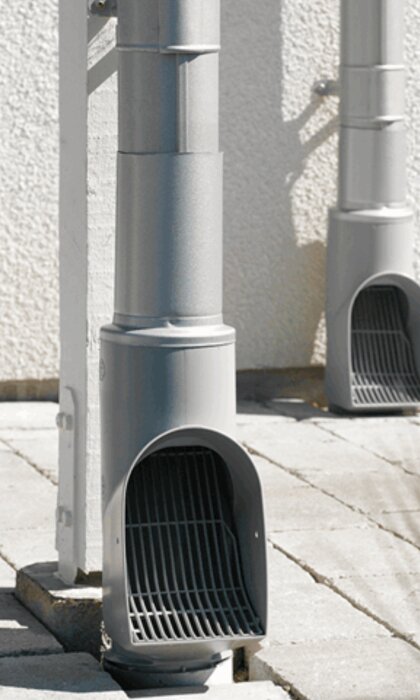 Två grå ventilationsrör med skyddsgaller vid husvägg, på betongplattor i dagsljus.