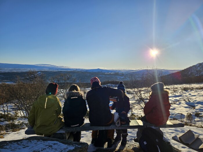 Personer sitter på bänk i snöig terräng, njuter av solen och utsikten över ett vinterlandskap.