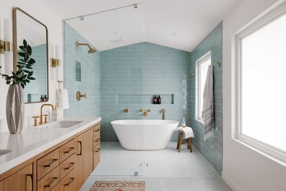 Modernt badrum, blå kakel, fristående badkar, träskåp, guldarmaturer, gröna växter, fönster, stilrent.