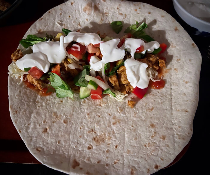 En öppen tortilla med sallad, tomat, kryddig kyckling, och gräddfil, redo att rullas till wrap.