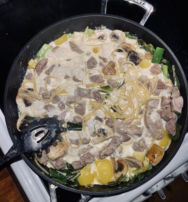 En stekpanna med gräddig pasta, kött, svamp, och grönsaker som tillagas på spisen.