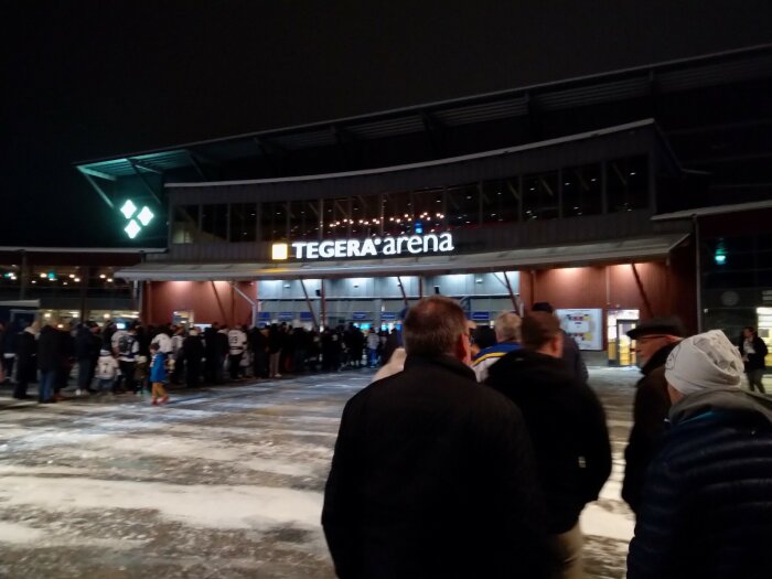 Kväll, folk utanför Tegera Arena, snötäckt mark, upplyst skylt, sannolikt före sportevenemang.