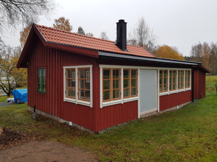 Rött trähus med vita knutar och fönster, skorsten, grönt gräs, mulen himmel, svensk landsbygd.