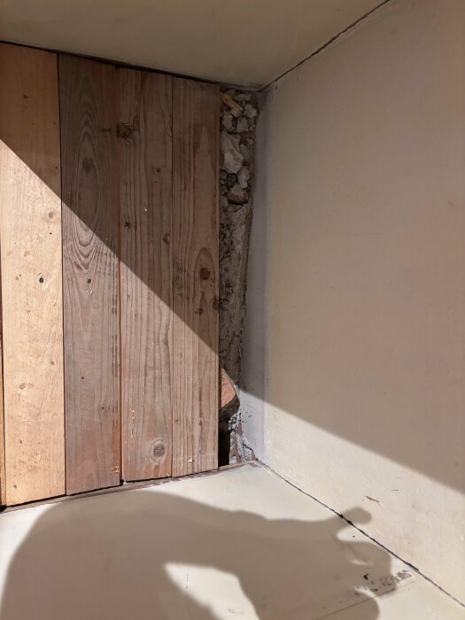 Hörn av rum med trägolv, vit vägg, skugga, reparationsbehov syns i betong och tegel.