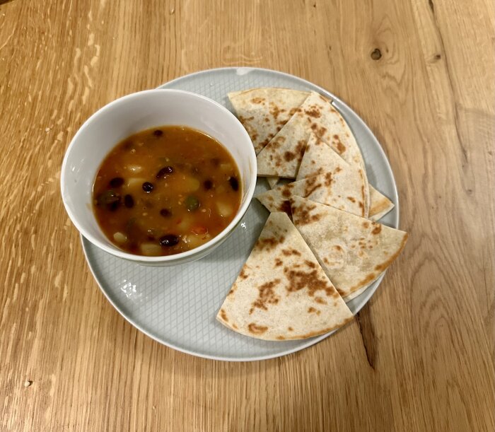 En vit skål med soppa, flankerad av fyra quesadillasegment på en tallrik, på ett träbord.