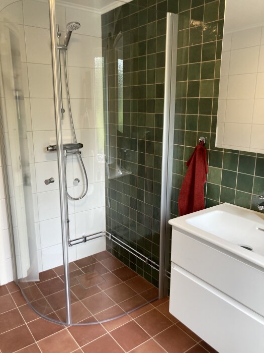 Badrum med duschhörna, vita skåp, gröna kakelväggar och terrakottafärgade klinkergolv.