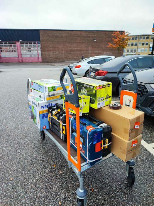 Handtruck lastad med verktyg och kartonger utanför en byggnad på parkeringsplats.