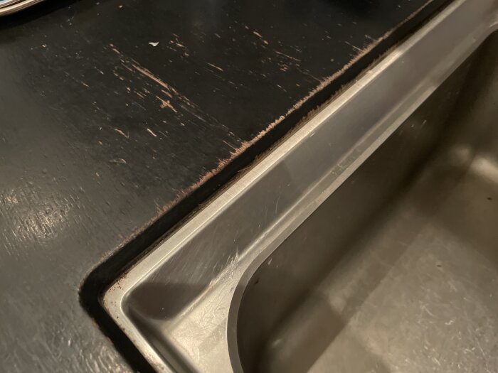 Närbild på slitet svart bord och rostfri stålkant, med synliga skador och repor.