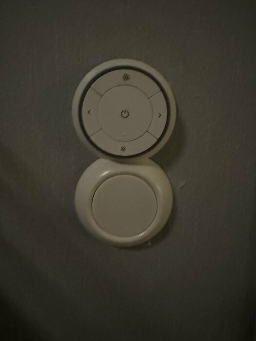 Vit väggmonterad enhet, möjlig termostat, knappar, rund design, mörk bakgrund.