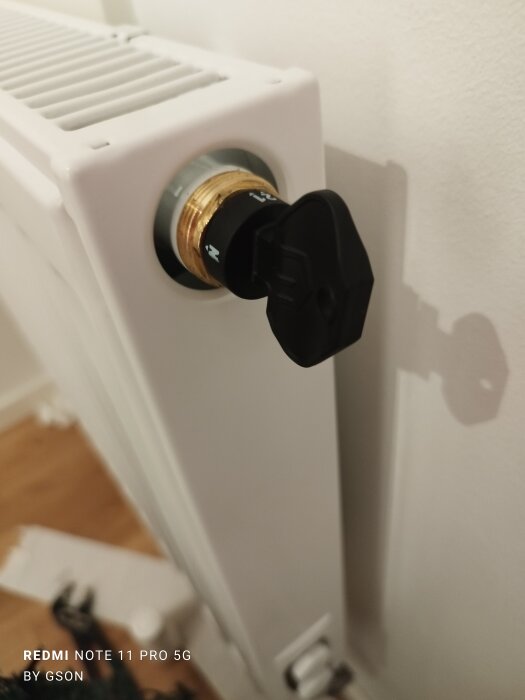 Termostatventil på vit radiator, justeringsratt, skugga på vägg, del av rum synlig.