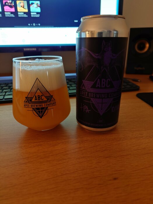 Ölglas med skum och burk på bord framför dataskärm och högtalare. Apex Brewing Company logotyp synlig.