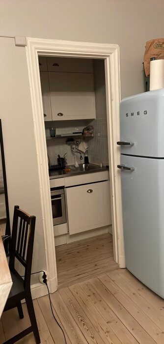 Kök med vit inredning, SMEG-kylskåp, trägolv, dörröppning från mörkt rum.