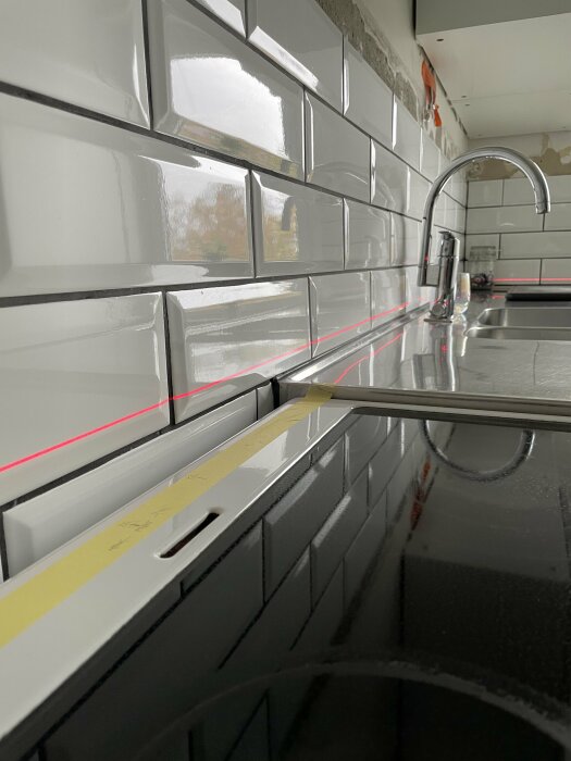 Kök under renovering, kakelvägg, vattenkran, lasernivå med röd linje, reflekterande arbetsbänk.