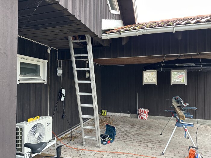 Carport med stege, värmepump, arbetsbänk och verktyg; tecken på installations- eller reparationsarbete pågår.