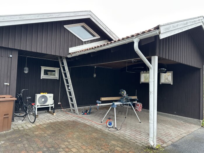 Carport med verktyg, stege, cykel och värmepump vid trähus. Arbetsbänk med såg utomhus. Cloudy day.