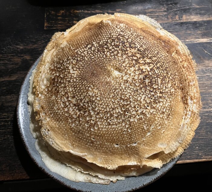 En hög nygräddade pannkakor på ett fat, brun-gyllene färg, bubblig yta, mörk bakgrund.