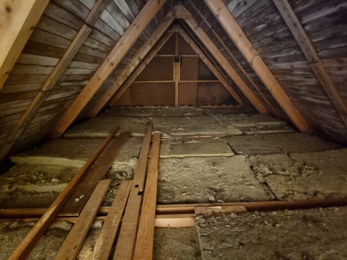 Gammalt vindsvåningsrum med träbalkar, isolering, och osorterade brädor. Dammtäckt, opolerat, och mörkt.