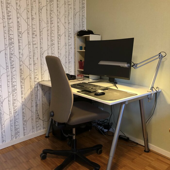 Ett kontorshörn med skrivbord, datorskärm, tangentbord, mus, stol och bordslampa. Trägolv, björkträdsmönstrad tapet.