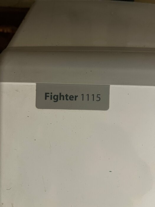 Etikett med text "Fighter 1115" på en vit yta med smutsfläckar, möjligen en apparat eller maskindel.