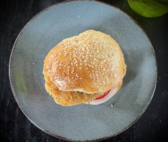 Sesambeströdd hamburgerbröd på en grå keramiktallrik mot en mörk bakgrund.