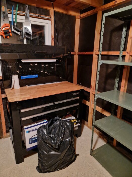 Verkstadsrum med arbetsbänk, tomma hyllor, verktygstavla, sopsäck och några verktyg.