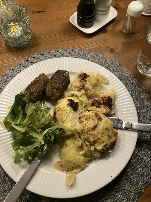 Middag med potatisgratäng, köttbullar, grönsallad på tallrik, vattenglas, salt- och pepparkvarnar i bakgrunden.