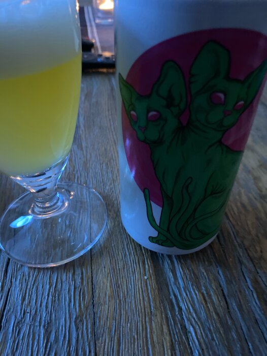 Glas med gul dryck intill mugg med gröna katter på träyta. Dimmigt och mysigt ljus.