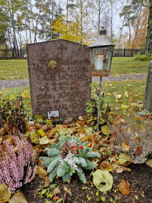 Gravsten omgiven av höstlöv, blommor och en tänd lykta på en kyrkogård. Lugn, höstig atmosfär.