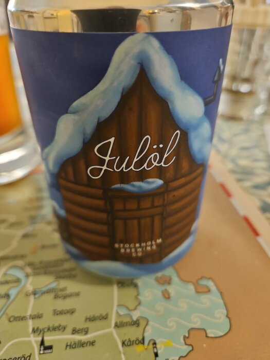 Ett ölglas med julmotiv och texten "Julöl" över en stiliserad bild av en stuga med snö.