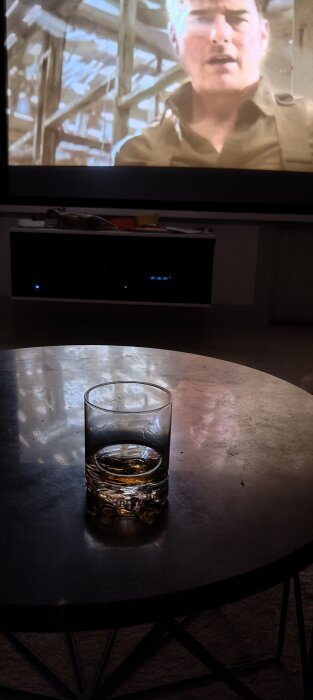 Vardagsrum med TV, person på skärm, whiskeyglas på soffbord, mörk belysning, hemtrevlig stämning.