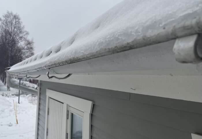 Snötäckt hustak i förgrund, is formationer på hängrännan, kyligt vinterlandskap, grå himmel.