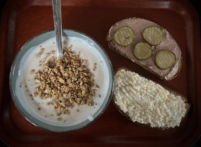 Yoghurt med granola, smörgåsar med leverpastej och inlagda gurkor samt keso på brunt träbricka.