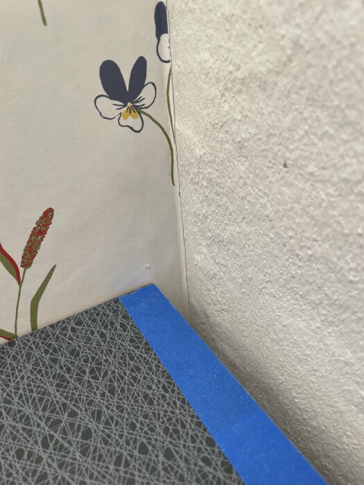 Blomtapetmönster, blått maskeringstejp, grått golv med geometriska mönster, hörn av ett rum.