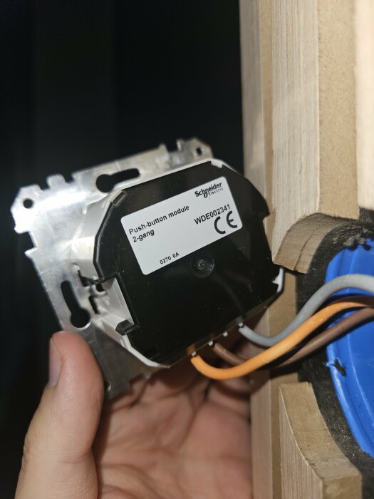 Hand håller en elektrisk tryckknappmodul nära vägg, kabelanslutning synlig, installationsprocess, Schneider Electric.