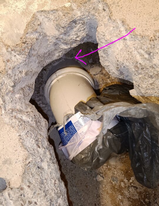 Rör synligt i brutet betonggolv, plastpåse och skräp blockerar; underhåll eller reparation eventuellt pågående.