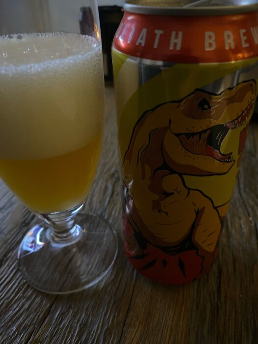 Ett glas med öl och en färgglad ölburk med en tecknad dinosaurie på en träyta.