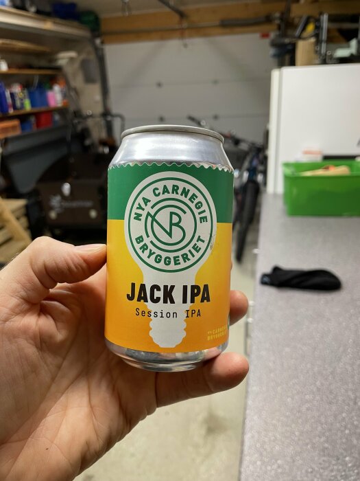 En hand håller en ölburk med texten "JACK IPA Session IPA" mot en oskarp garagebakgrund.