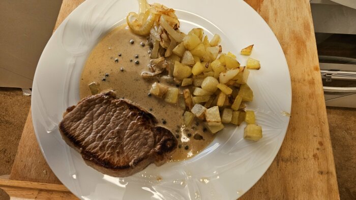 Stekt biff med gräddsås, kuber av stekt potatis, lök, svartpepparkorn på vit tallrik.