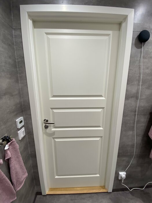 En vit panelad dörr med handtag, grå kakelvägg, golvstående lampa, handdukar och strömkontakter.