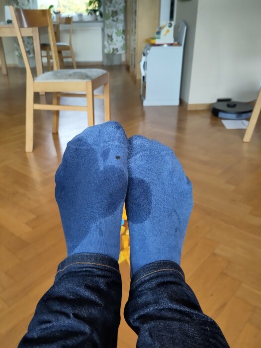 Fötter i blåa strumpor, jeans, inomhusmiljö med stolar, bord, kyl och robotdammsugare. Strumporna har ett hål.